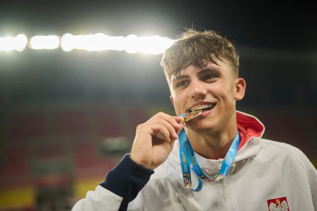 Nikodem Dymiński ze złotym medalem EYOF Młody sportowiec w białym dresie z godłem Polski na piersi trzyma medale na niebieskiej wstążce i uśmiecha się, lekko gryząc medal na tle rozświetlonego stadionu.
