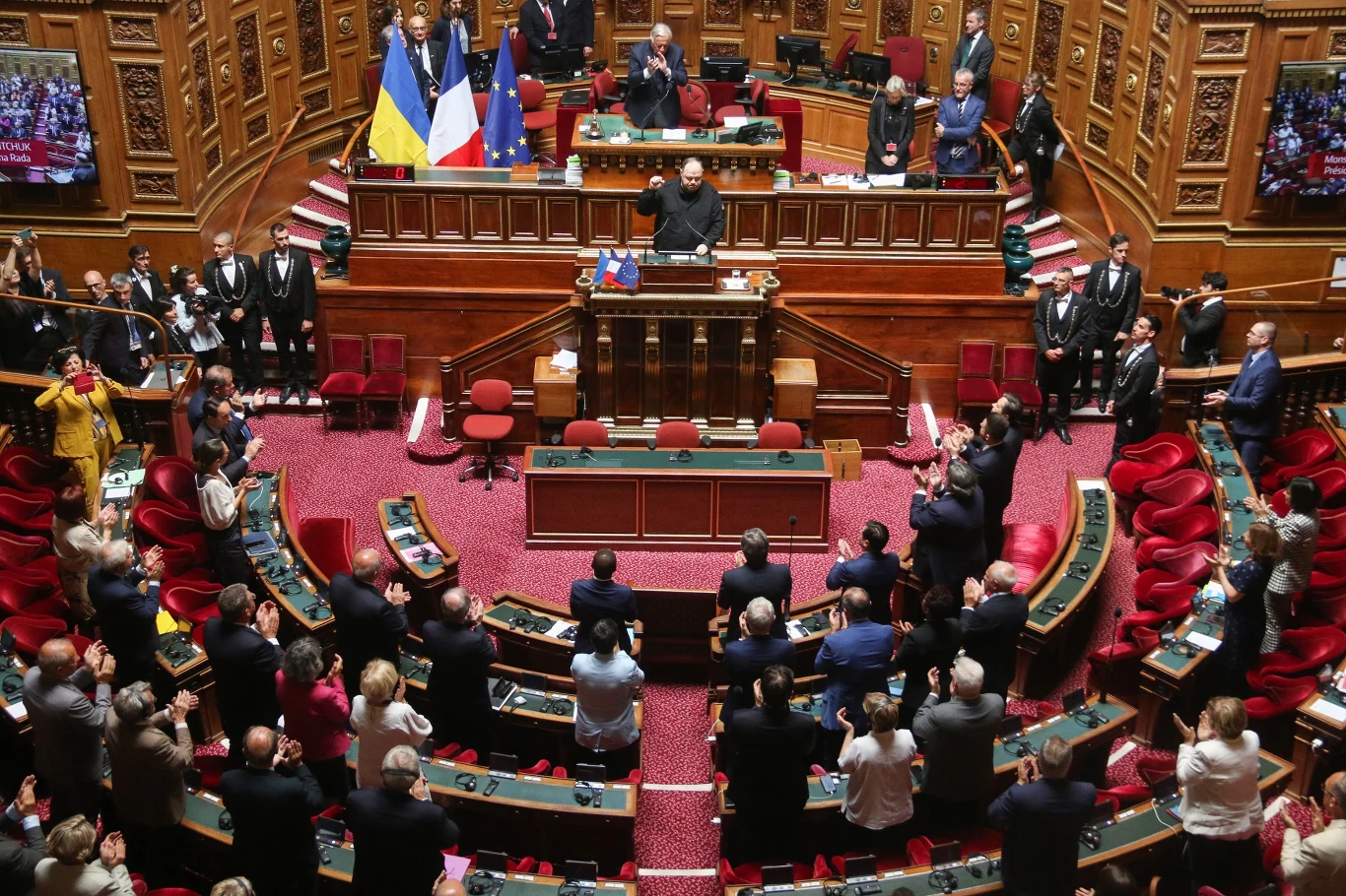Ukraiński parlament Sala parlamentarna podczas oficjalnego przemówienia, liczne osoby ubrane elegancko, widoczne flagi Ukrainy, Francji oraz Unii Europejskiej za mównicą, zgromadzeni parlamentarzyści oraz ochrona.