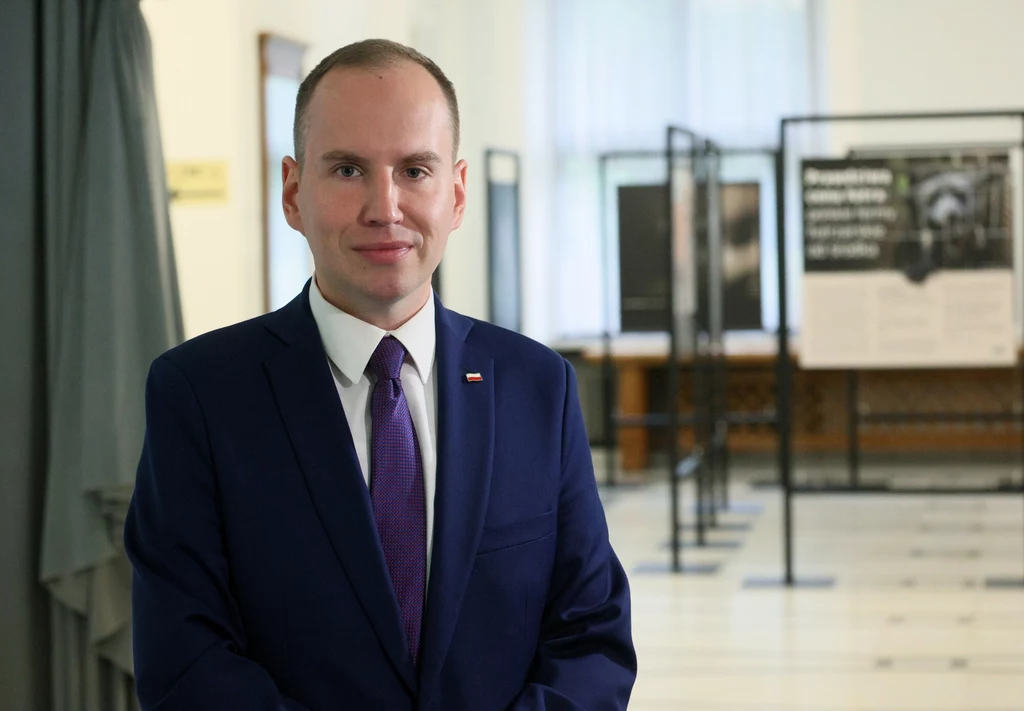 Elegancko ubrany mężczyzna w granatowym garniturze z fioletowym krawatem stoi w jasnym, oficjalnym wnętrzu z tablicami informacyjnymi w tle.