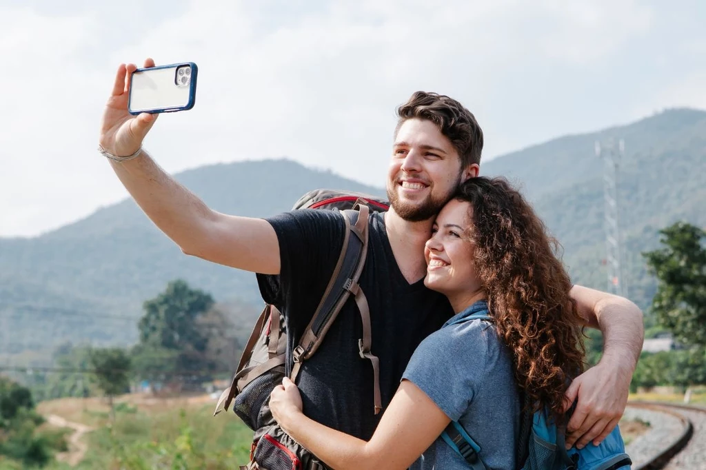 Para turystów z plecakami obejmuje się i uśmiecha wykonując wspólne selfie na tle górskiego krajobrazu.
