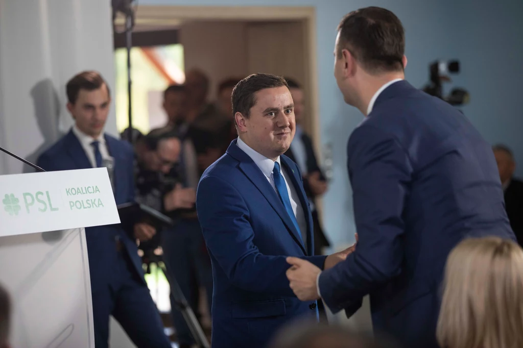 Dwóch mężczyzn w garniturach wita się uściskem dłoni podczas oficjalnego wydarzenia PSL Koalicja Polska, w tle stoją inni uczestnicy i aparat fotograficzny skierowany na scenę.