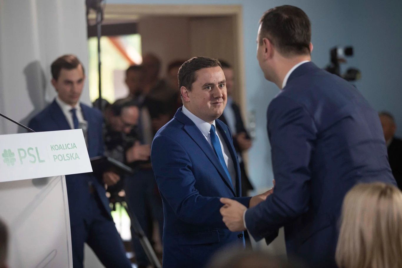Dwóch mężczyzn w garniturach wita się uściskem dłoni podczas oficjalnego wydarzenia PSL Koalicja Polska, w tle stoją inni uczestnicy i aparat fotograficzny skierowany na scenę.