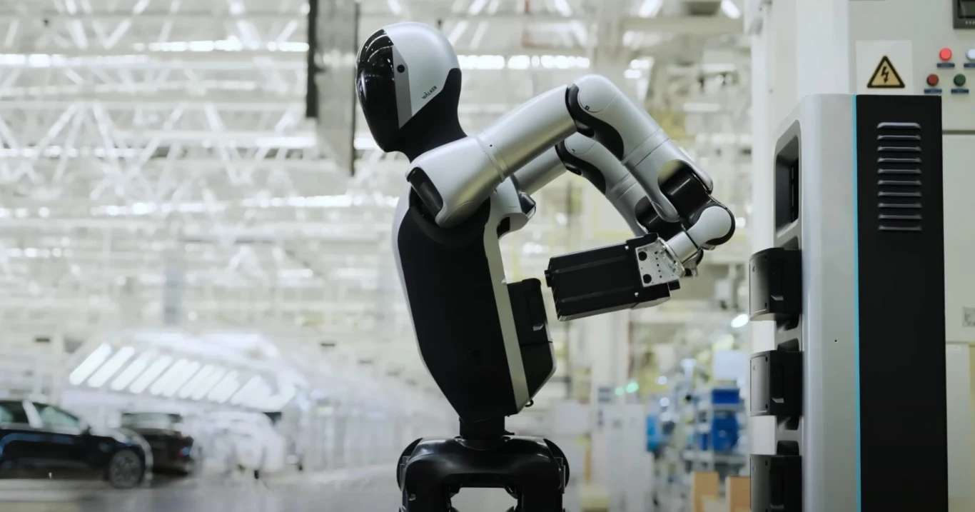 Walker S2 to robot, który sam wymienia sobie baterię i pracuje non stop Robot humanoidalny wykonujący czynności przy panelu sterowniczym w nowoczesnej fabryce, z widocznym w tle samochodem i industrialnym otoczeniem.