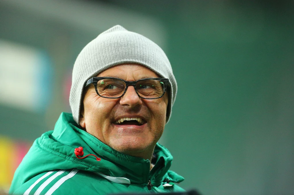 Mężczyzna w okularach, ubrany w zieloną kurtkę sportową i szarą czapkę, uśmiecha się szeroko, tło rozmazane w zielonych odcieniach.