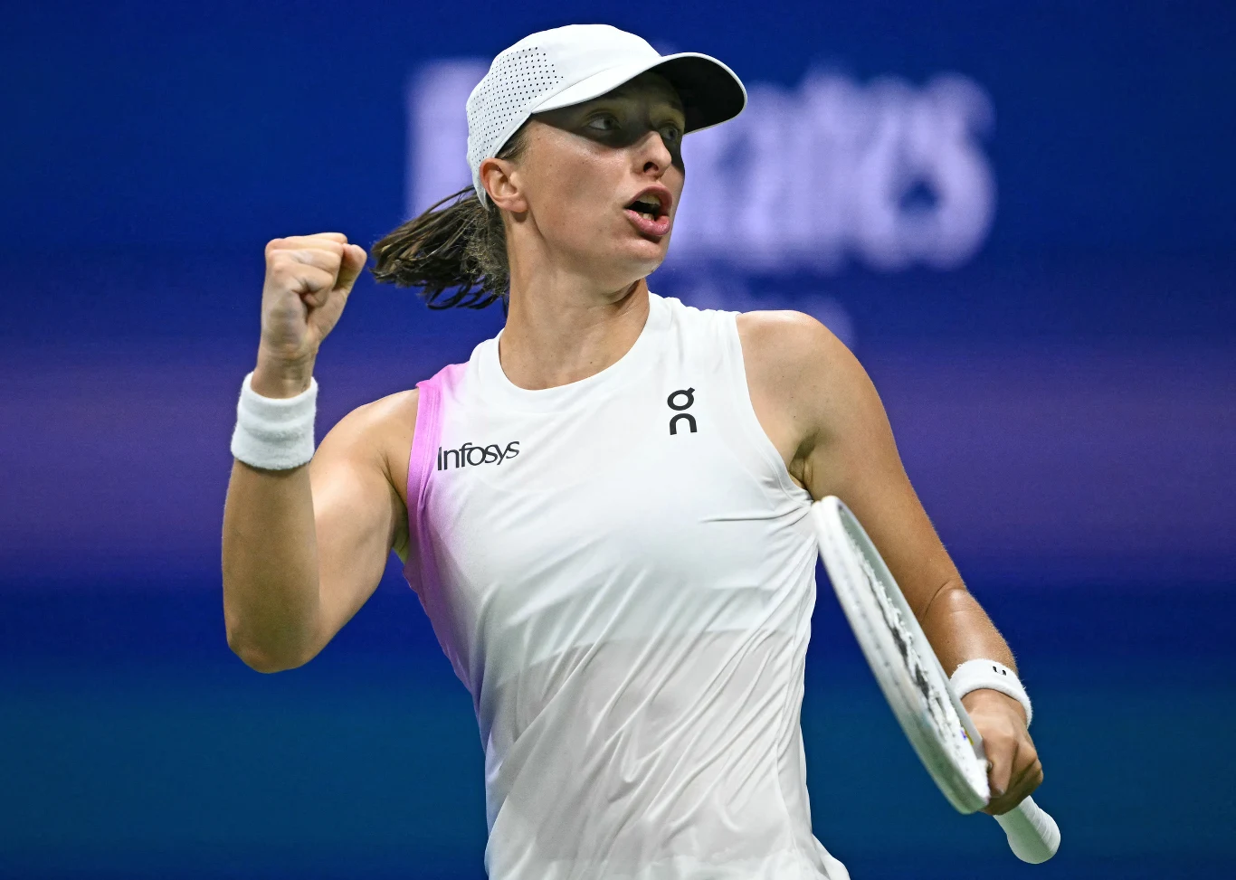 Iga Świątek Tenisistka ubrana na biało w czapce z daszkiem świętuje zwycięski punkt, zaciskając pięść i trzymając rakietę sportową w lewej ręce, tło rozmyte z niebieskimi barwami kortu.