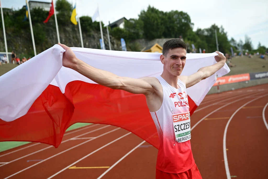 Mikołaj Szczęsny Lekkoatleta w stroju reprezentacji Polski trzyma dużą polską flagę, stojąc na bieżni stadionu lekkoatletycznego; w tle widoczne są trybuny, flagi i teren rekreacyjny.