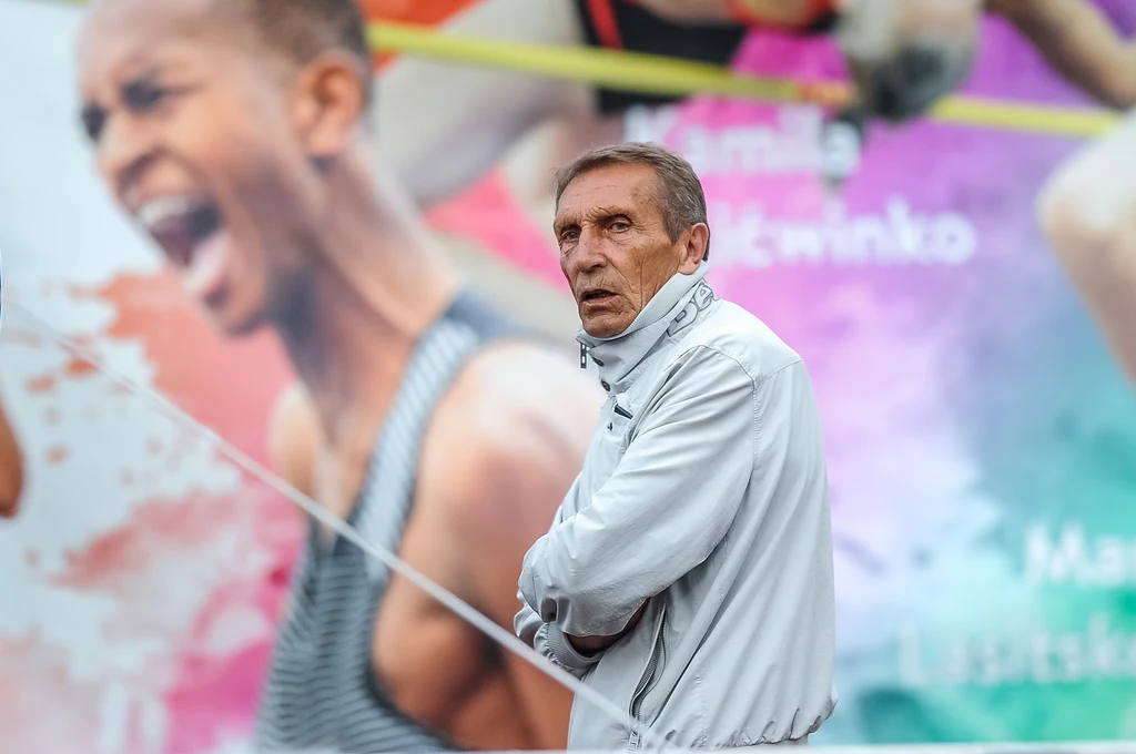 Włodzimierz Michalski Mężczyzna w sportowej kurtce na tle kolorowego baneru z wizerunkami sportowców, wyrażających emocje podczas rywalizacji.