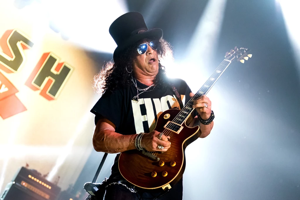 Slash w akcji: czarne loki, okulary przeciwsłoneczne, cylinder na głowie i gryf gitary zadarty w górę Slash w akcji: czarne loki, okulary przeciwsłoneczne, cylinder na głowie i gryf gitary zadarty w górę