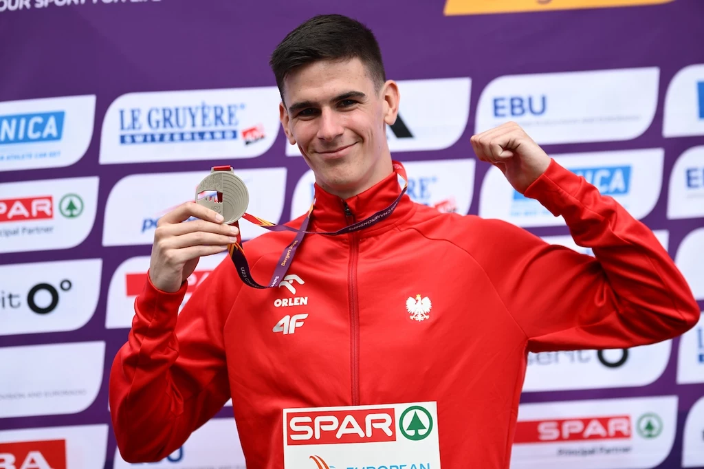 Mikołaj Szczęsny ze srebrnym medalem MME Młody sportowiec w czerwonym dresie z polskim godłem prezentuje srebrny medal, uśmiechając się i unosząc zaciśniętą pięść w geście triumfu, na tle tablicy sponsorów i logotypów.