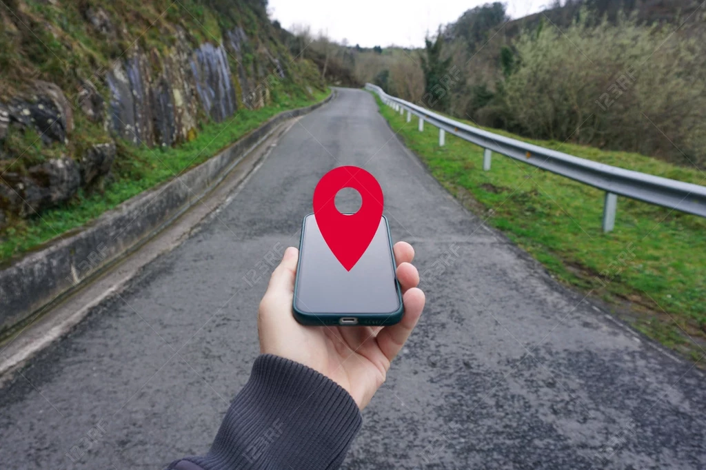 Ręka trzymająca smartfon z wyświetlonym czerwonym znacznikiem lokalizacji na tle asfaltowej drogi prowadzącej przez zalesiony teren.
