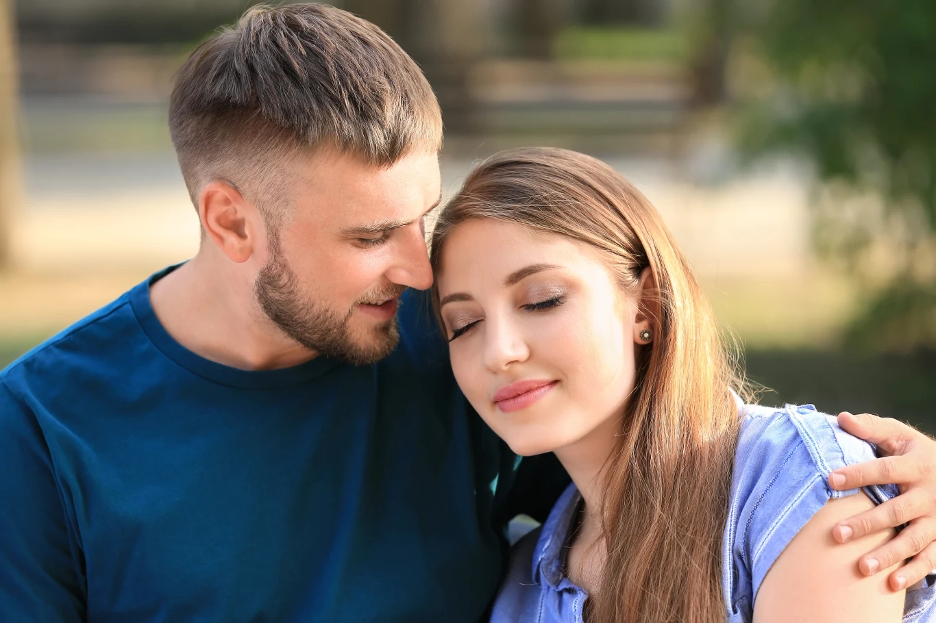 Mężczyzna obejmuje młodą kobietę, która z zamkniętymi oczami opiera głowę na jego ramieniu, oboje wyglądają na spokojnych i zrelaksowanych, w tle rozmyte drzewa i ławka w parku