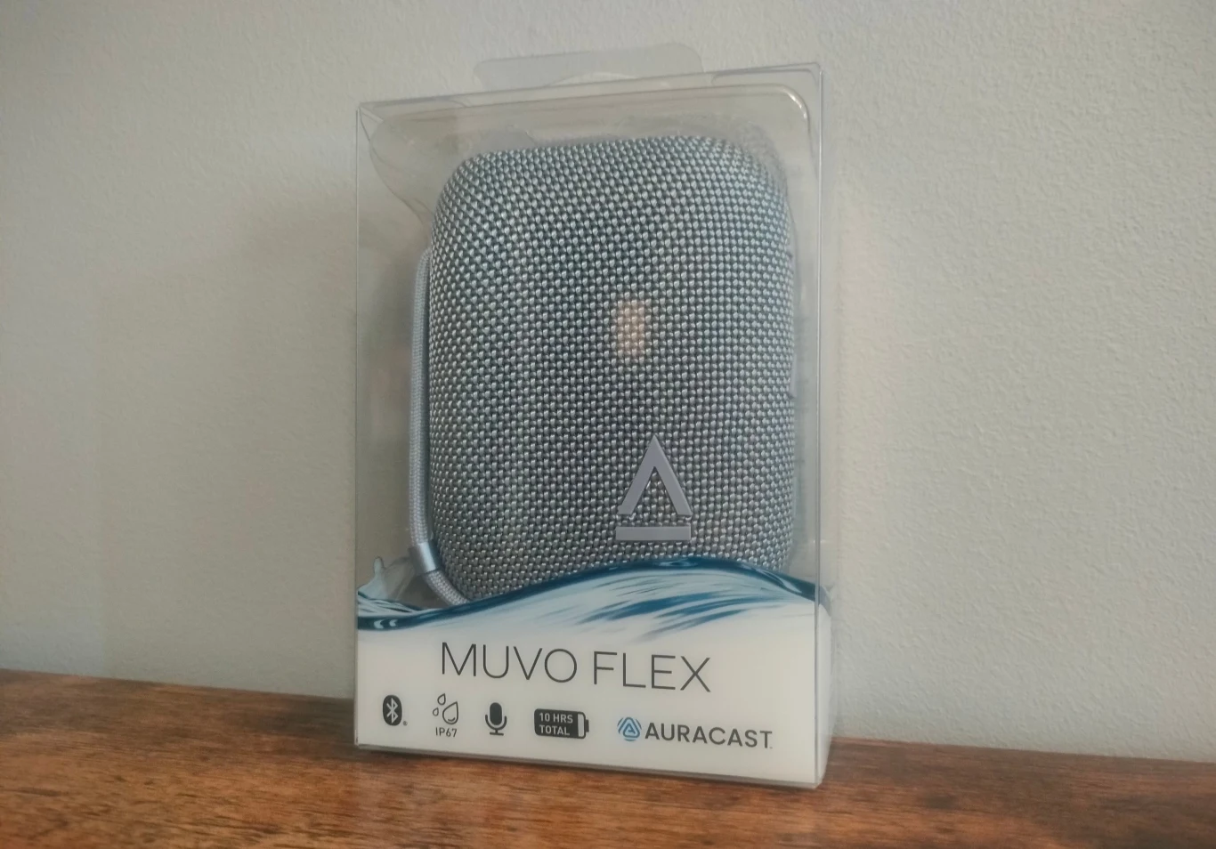 Creative Muvo Flex Głośnik Bluetooth MUVO Flex w oryginalnym, przezroczystym opakowaniu stojącym na drewnianej powierzchni, widoczne są oznaczenia producenta i funkcje urządzenia, jak Bluetooth, mikrofon oraz logo Auracast.
