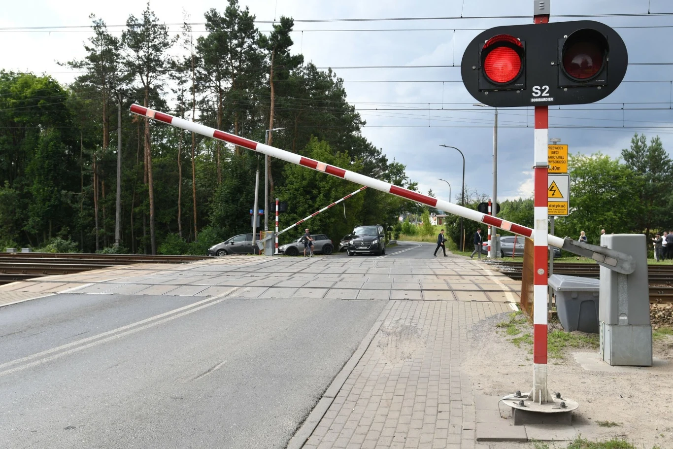 Szlaban opuszczony na przejeździe kolejowym, czerwone światła sygnalizacyjne i kilka czekających samochodów na drodze przed torami, w tle widoczne drzewa i słupy trakcyjne.
