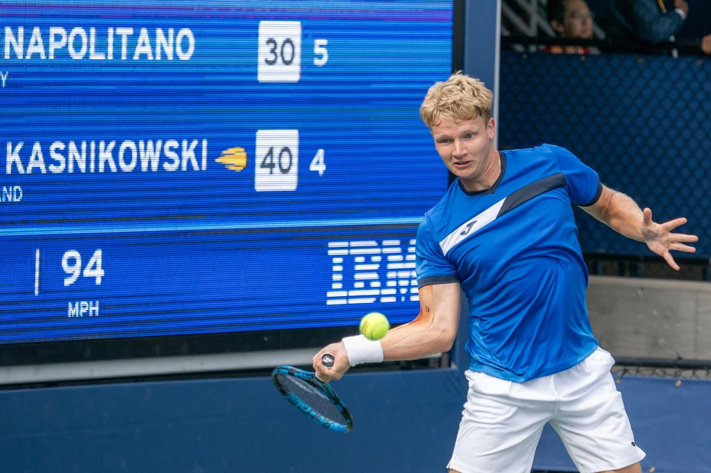 Kaśnikowski ma za sobą debiut w US Open Zawodnik uderza piłkę tenisową rakietą podczas meczu, w tle elektroniczna tablica z wynikami i czasem, na której widoczne są nazwiska graczy oraz prędkość serwu.