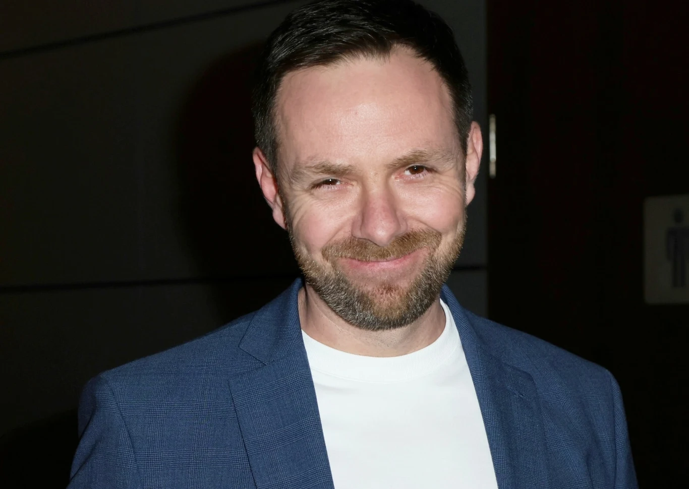 Adam "Łona" Zieliński Mężczyzna z krótkimi, ciemnymi włosami i brodą ubrany w niebieską marynarkę oraz biały t-shirt stoi na tle jednolitej, ciemnej ściany, uśmiecha się lekko do obiektywu.