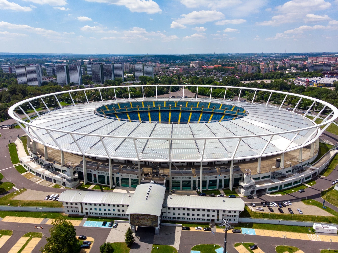Nowoczesny stadion piłkarski otoczony miejską zabudową, widoczny z lotu ptaka, z wyróżniającą się białą konstrukcją dachu oraz wysokimi trybunami, wokół stadionu zaparkowane samochody, w tle panorama miasta.
