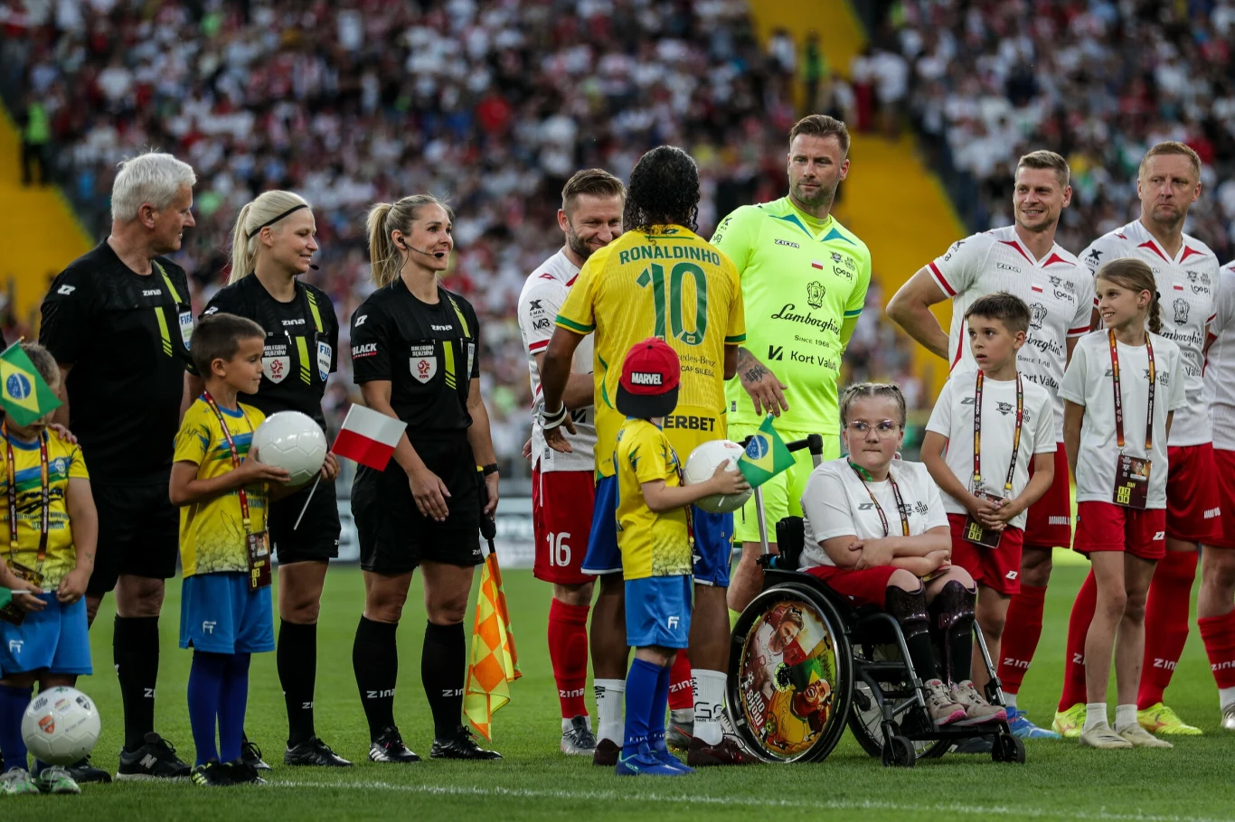 Piłkarze obu drużyn stojący na murawie boiska wraz z dziećmi, w tym dzieckiem na wózku inwalidzkim, towarzyszą im sędziowie oraz wolontariusze, w tle wypełnione kibicami trybuny stadionu.