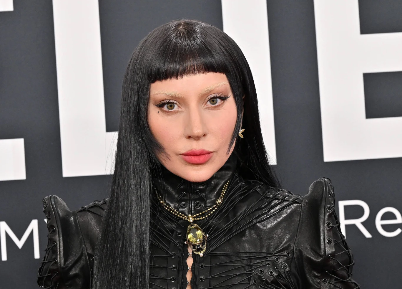 Lady Gaga wywróciła się podczas koncertu Kobieta o długich czarnych włosach z prostą grzywką, ubrana w czarną skórzaną kurtkę z ozdobnym naszyjnikiem, stoi na tle ciemnej planszy z dużymi, białymi literami.
