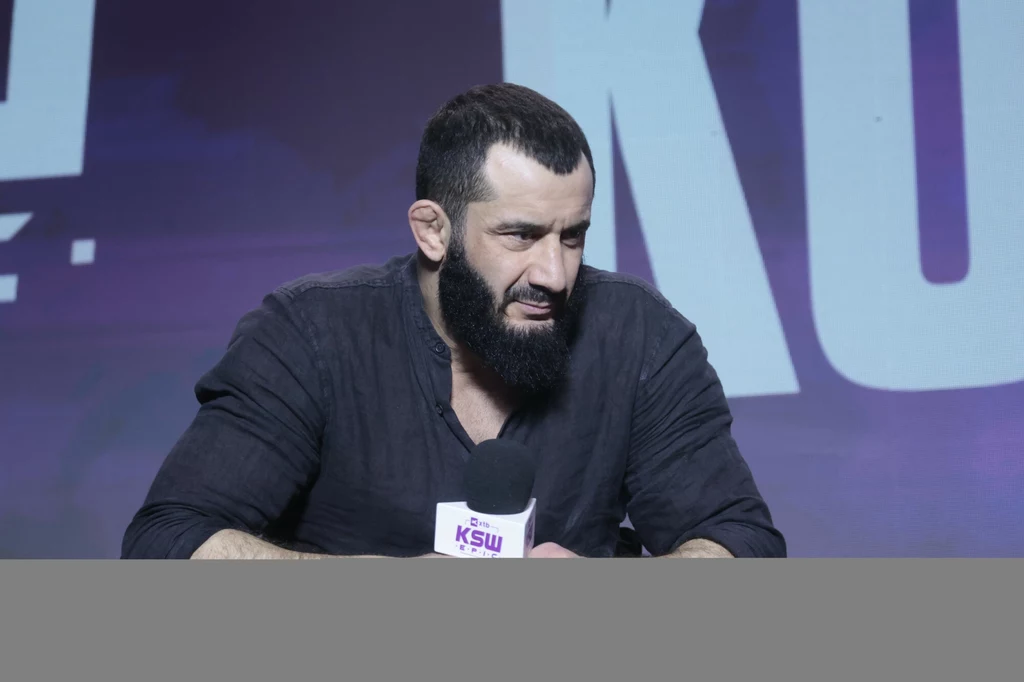 Mamed Chalidow Mężczyzna z ciemną brodą w czarnej koszuli siedzi przy stole, trzyma w dłoni mikrofon z logo KSW, w tle rozmyte duże litery na fioletowym tle.