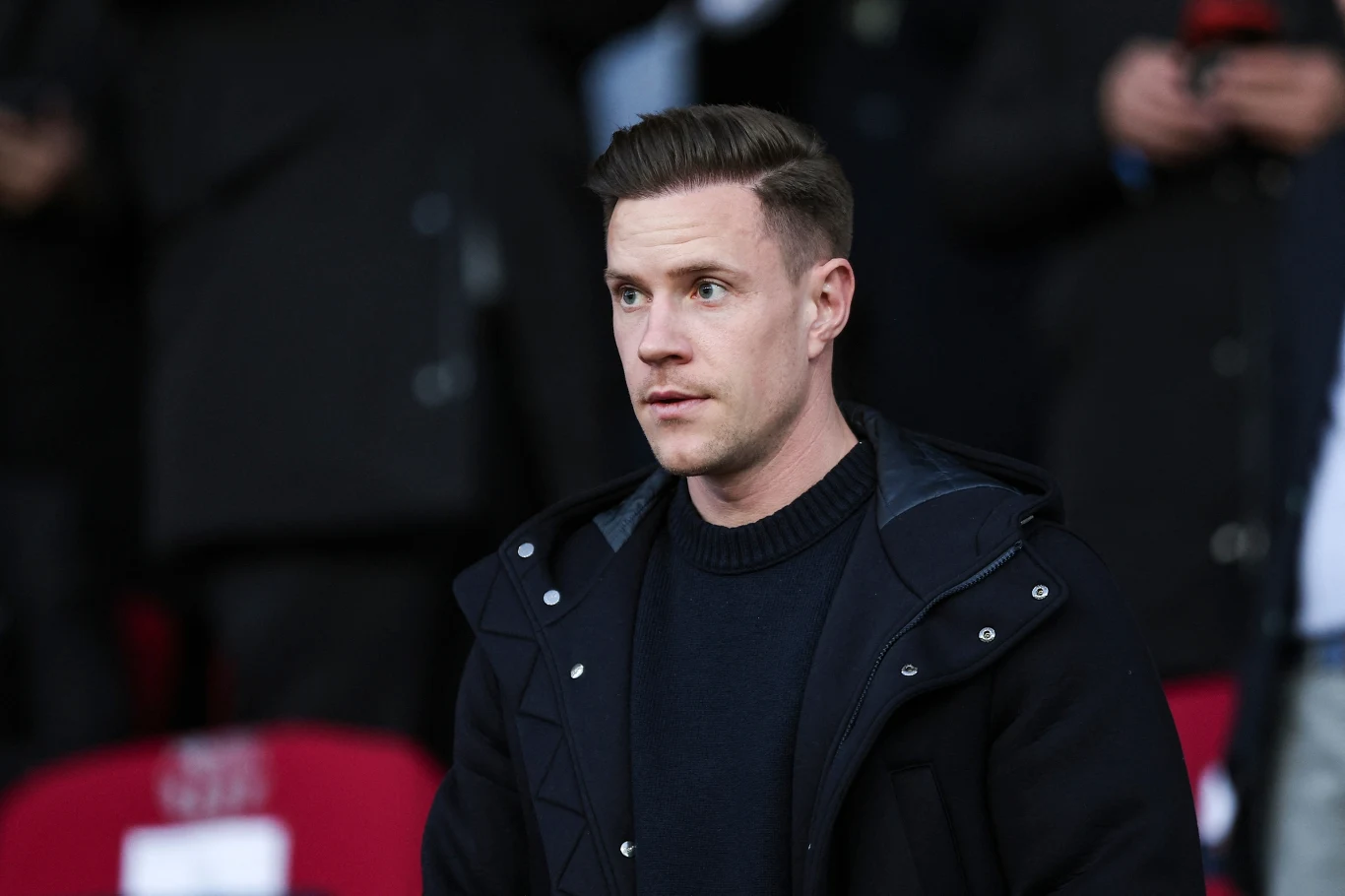 Marc-Andre ter Stegen Mężczyzna o krótkich, zaczesanych do góry włosach, ubrany w ciemny sweter i kurtkę, znajduje się na trybunach, w tle widoczni rozmyci ludzie i czerwone krzesełka.