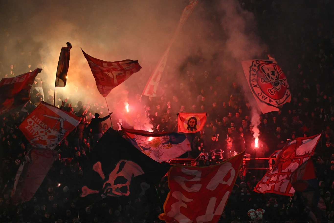 Kibice Crveny zvezdy Belgrad Tłum kibiców na stadionie w ciemności trzyma czerwone flagi oraz race pirotechniczne, wokół unosi się dym, a na flagach widać czaszki i klubowe symbole.