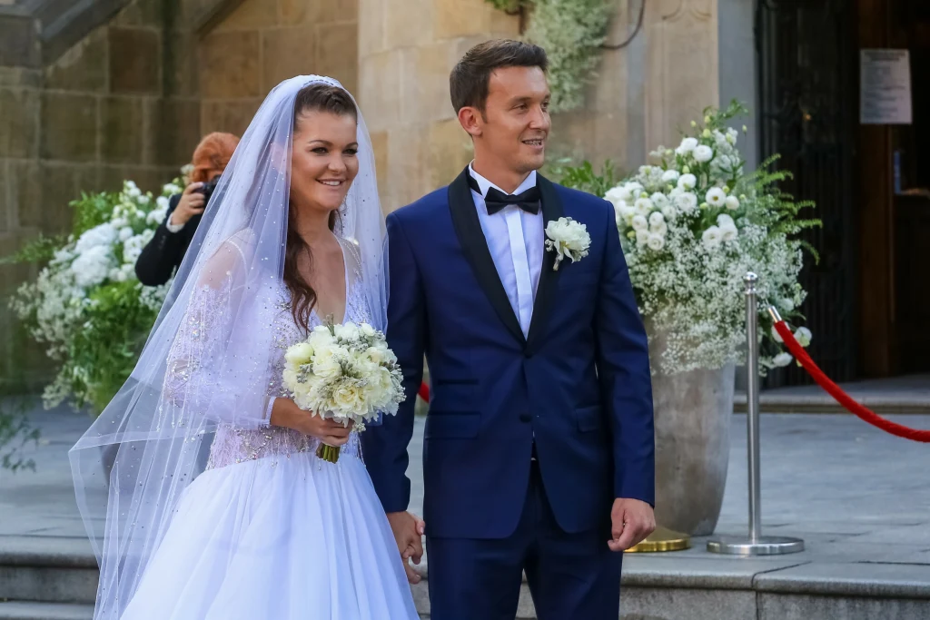 Agnieszka Radwańska i Dawid Celt Para młoda wychodzi z kościoła, ubrana w eleganckie stroje ślubne, panna młoda trzyma bukiet białych kwiatów, w tle kwiatowe dekoracje i fotograf robiący zdjęcia.