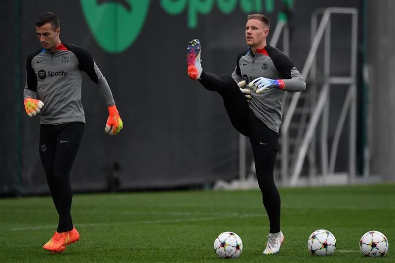 Dwóch bramkarzy drużyny piłkarskiej podczas treningu na murawie, jeden z nich wykonuje rozgrzewkowe kopnięcie, a drugi trzyma piłkę w dłoniach, przed nimi leżą futbolówki. Obaj są ubrani w jednolite stroje treningowe w barwach klubu FC Barcelona.