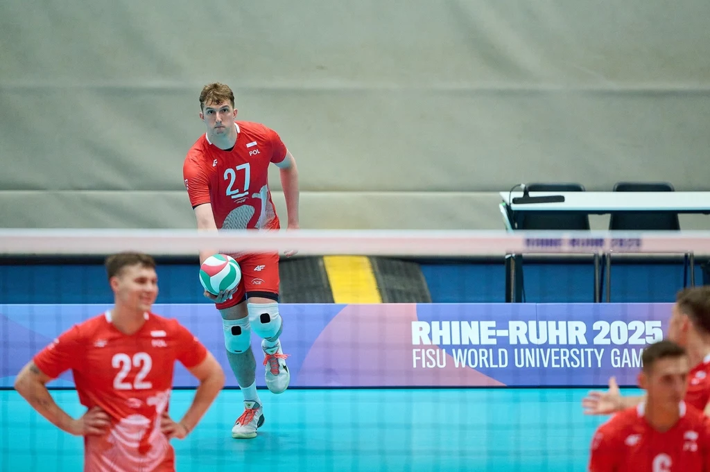 Michał Gierżot, kapitan polskiej kadry na Uniwersjadę Zawodnik w czerwonym stroju podczas serwowania piłki siatkowej na hali, w tle widoczni inni sportowcy oraz banner z informacją o wydarzeniu Rhine-Ruhr 2025 FISU World University Games.