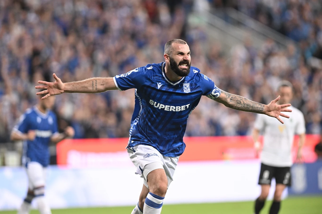 Lech Poznań pewnie pokonał Breidablik Piłkarz w niebieskiej koszulce drużyny Lecha Poznań z wyciągniętymi rękami świętuje zdobycie bramki, w tle rozmazana publiczność oraz zawodnicy drużyny przeciwnej w białych strojach.