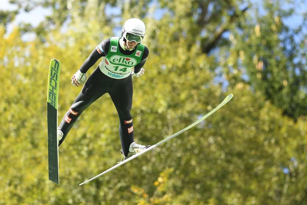 Skoczek narciarski w kombinezonie sportowym i kasku wykonuje skok na tle drzew, unosząc się w powietrzu z nartami rozstawionymi szeroko.