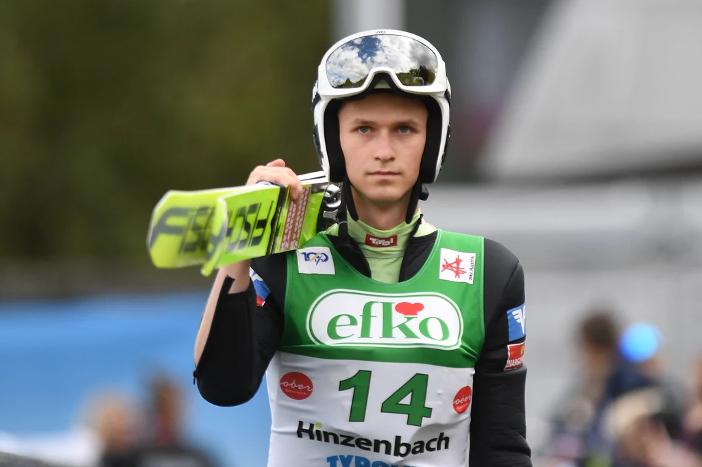 Zawodnik skoków narciarskich ubrany w strój sportowy oraz kask, trzymający narty na ramieniu, z numerem startowym 14 i widocznymi logotypami sponsorów na stroju.