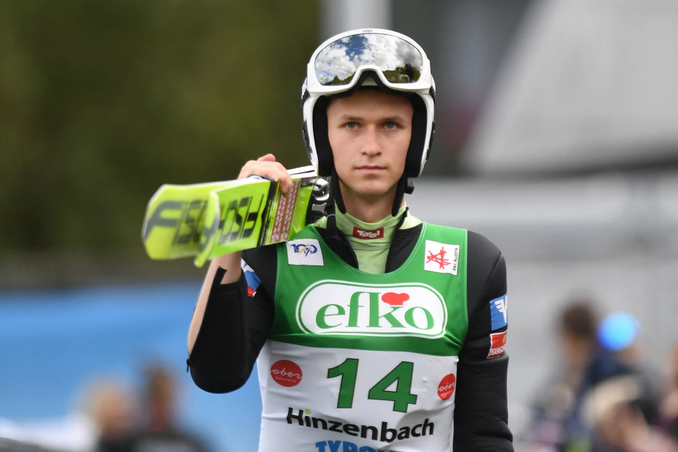 Zawodnik skoków narciarskich ubrany w strój sportowy oraz kask, trzymający narty na ramieniu, z numerem startowym 14 i widocznymi logotypami sponsorów na stroju.