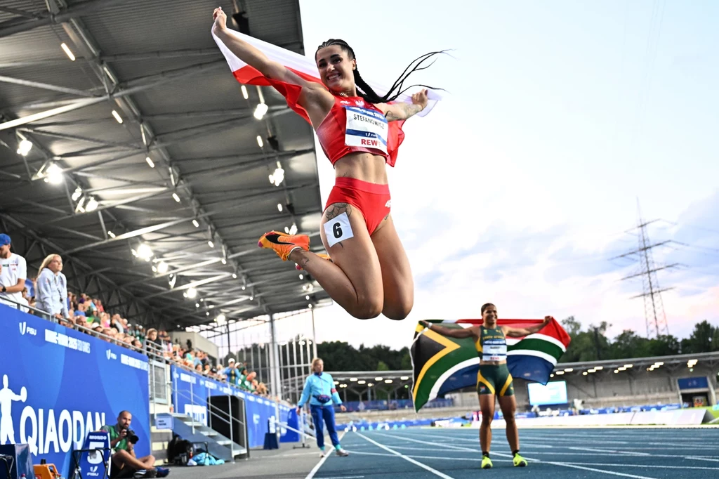 Magdalena Stefanowicz Polska lekkoatletka w czerwonym stroju sportowym skacze z radości po zwycięstwie, trzymając biało-czerwoną flagę, w tle zawodniczka z RPA z flagą swojego kraju oraz fragment bieżni stadionu lekkoatletycznego.