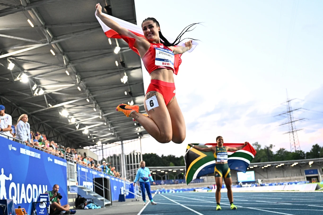 Polska lekkoatletka w czerwonym stroju sportowym skacze z radości po zwycięstwie, trzymając biało-czerwoną flagę, w tle zawodniczka z RPA z flagą swojego kraju oraz fragment bieżni stadionu lekkoatletycznego.
