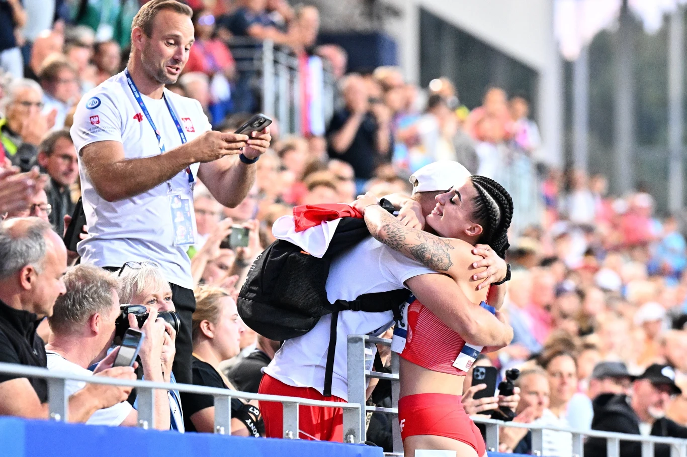 Sportsmenka w czerwonym stroju obejmuje mężczyznę z plecakiem, wokół stoją kibice i fotografują ten moment, klimat trybun jest pełen radości i emocji po sukcesie sportowym.
