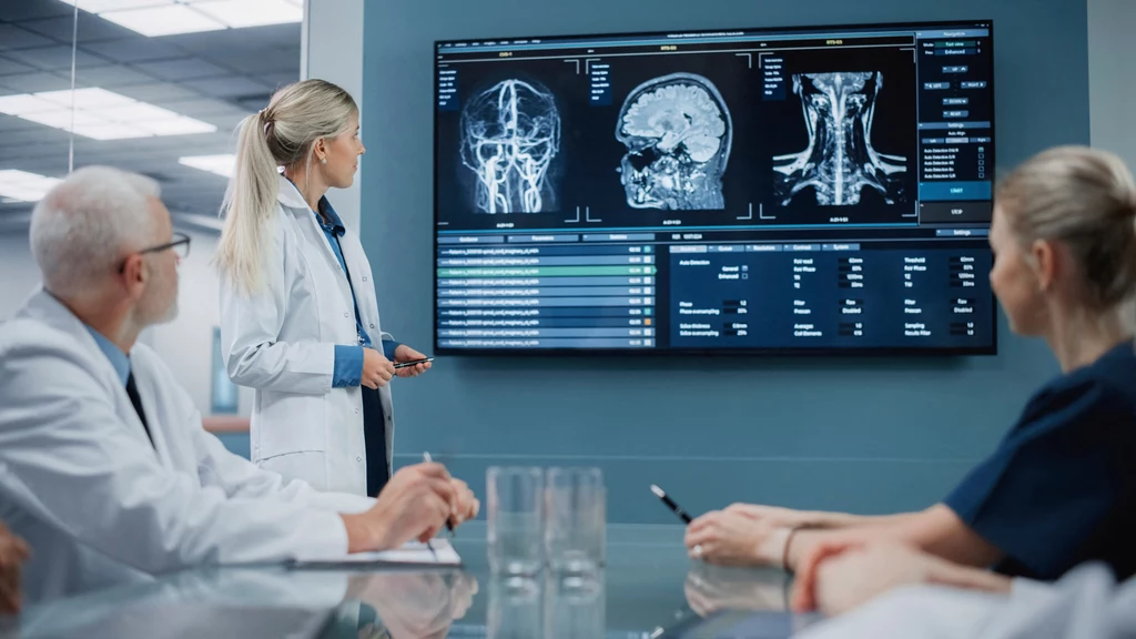 Rezonans magnetyczny to nieinwazyjna metoda diagnostyki obrazowej, która wykorzystuje silne pole magnetyczne i fale radiowe do uzyskiwania szczegółowych obrazów wnętrza ciała Lekarka prezentuje zespołowi medycznemu wyniki obrazowania mózgu i kręgosłupa wyświetlone na dużym ekranie, zespół skupiony na analizie zdjęć medycznych.