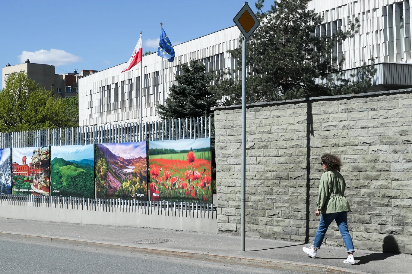Ambasada RP w Moskwie Budynek instytucji publicznej z ogrodzeniem ozdobionym dużymi obrazami krajobrazów, przed którym przechodzi kobieta ubrana w zieloną kurtkę i jeansy, widoczne flagi Polski i Unii Europejskiej.