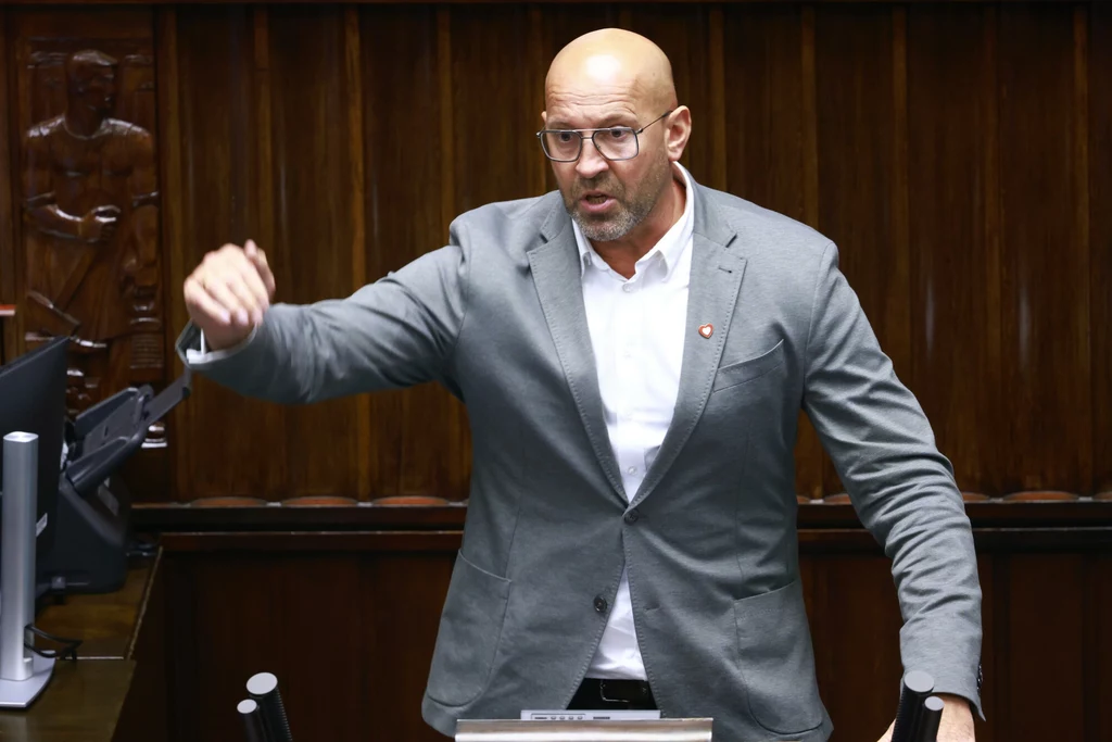 Jakub Rutnicki Mężczyzna w okularach i szarym garniturze przemawia energicznie przy mównicy w drewnianej sali, gestykulując ręką, w tle widoczne są rzeźbione elementy drewnianej boazerii.