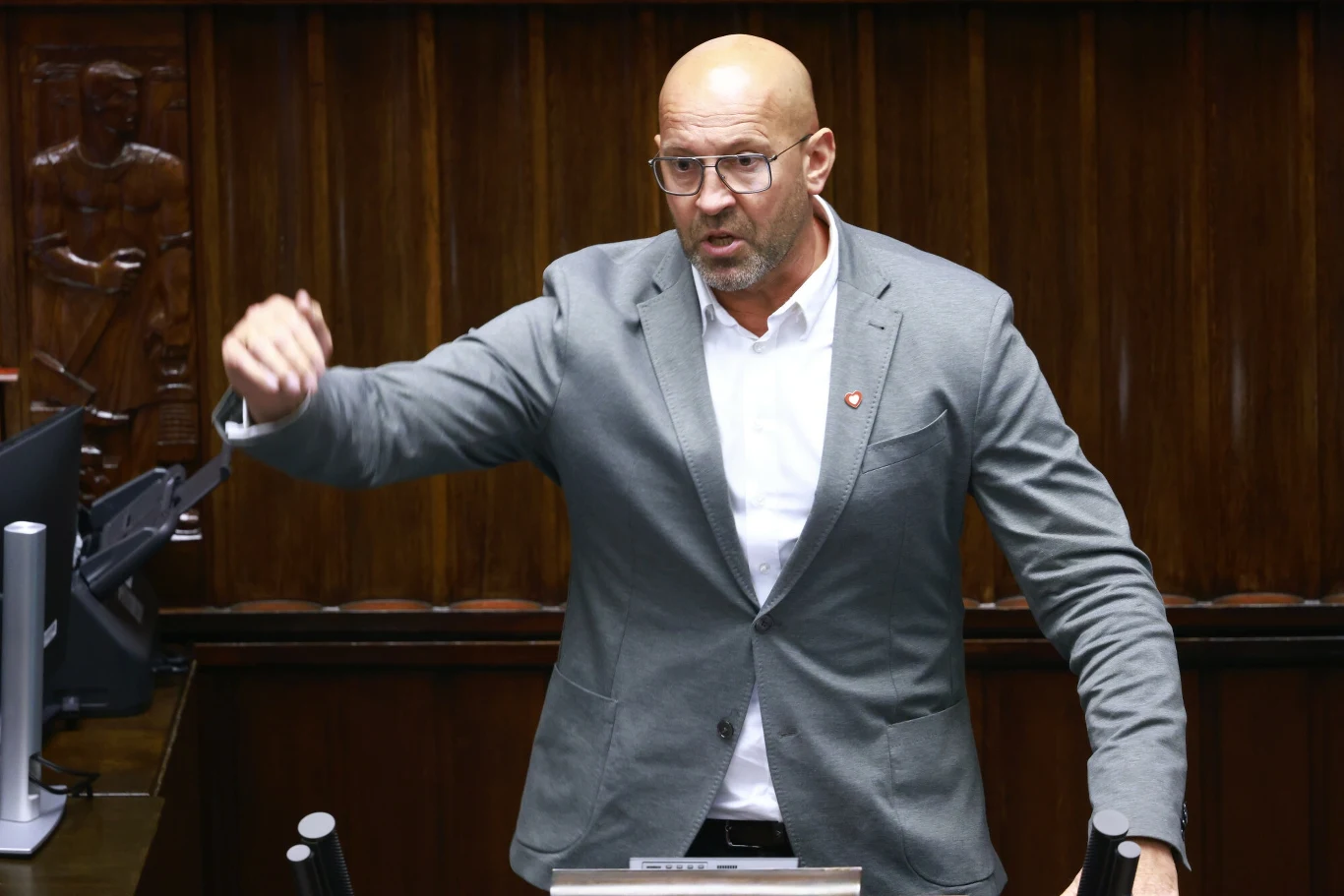 Mężczyzna w okularach i szarym garniturze przemawia energicznie przy mównicy w drewnianej sali, gestykulując ręką, w tle widoczne są rzeźbione elementy drewnianej boazerii.