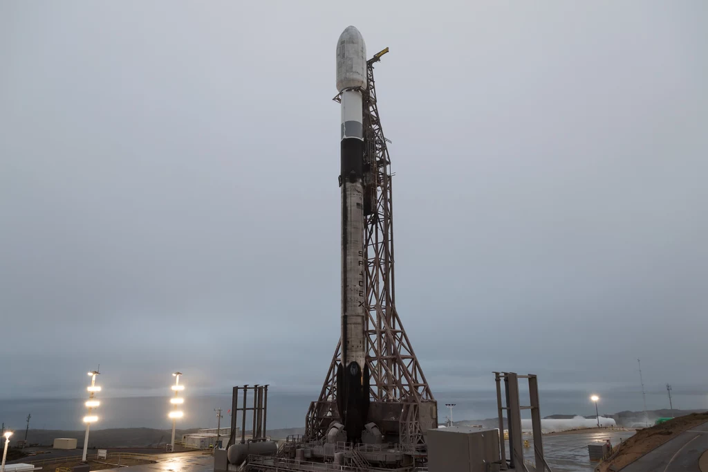 Rakieta Falcon 9, która wyniesie satelity TRACERS należące do NASA. Rakieta przygotowana do startu, stojąca na platformie startowej w otoczeniu infrastruktury technicznej, widoczne światła i pochmurne niebo w tle.