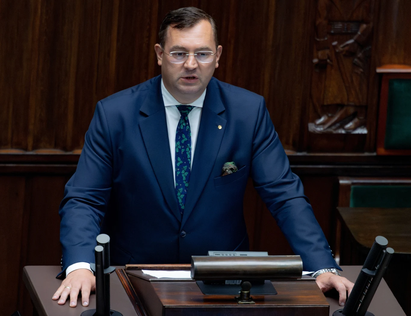 Stefan Krajewski zapowiedział ustawę, która ma ochronić gospodarstwa rolne. Mężczyzna w garniturze przemawia z mównicy sejmowej, w tle fragment dekoracji drewnianych i zielone obicie ław poselskich.