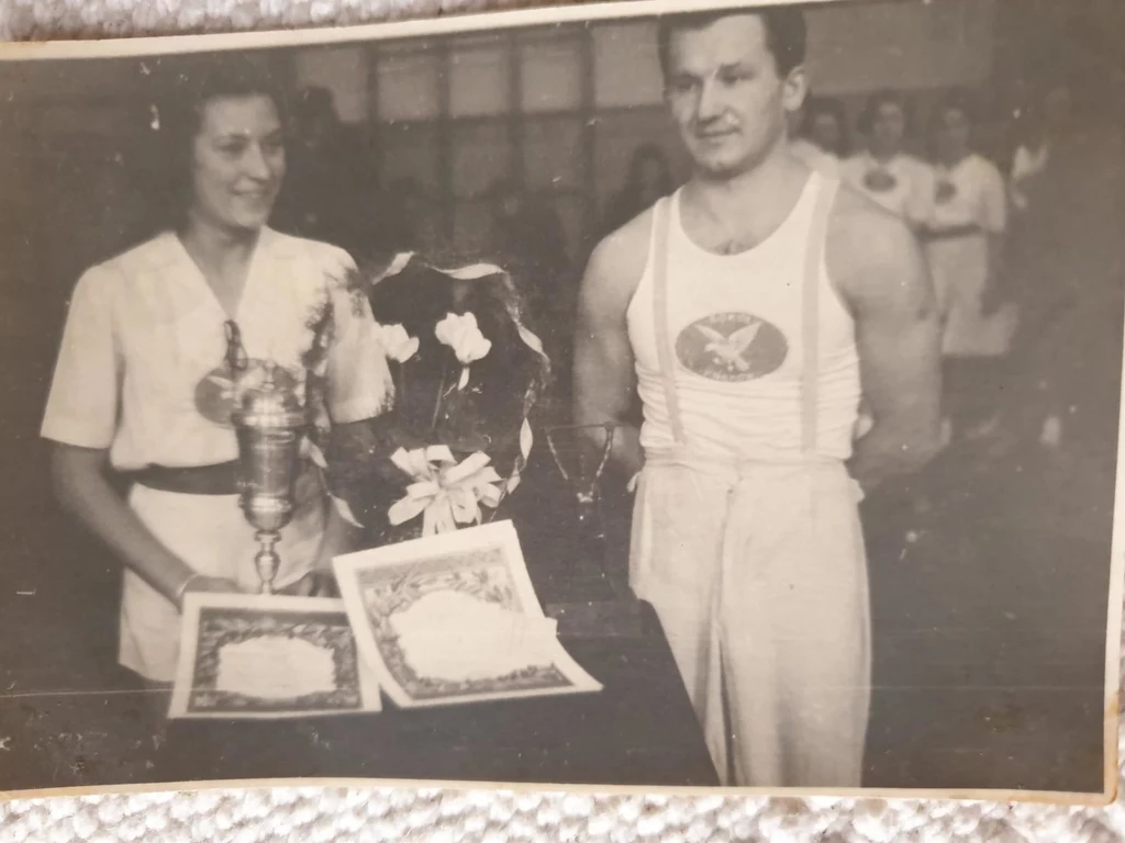 Pani Zosia z pucharem - zdjęcie z początkowego okresu Sokoła Kobieta w białym ubraniu i mężczyzna w stroju sportowym stoją obok siebie, na stole przed nimi znajdują się puchary, bukiet kwiatów oraz dwa dyplomy, w tle kilka osób w podobnych strojach sportowych.