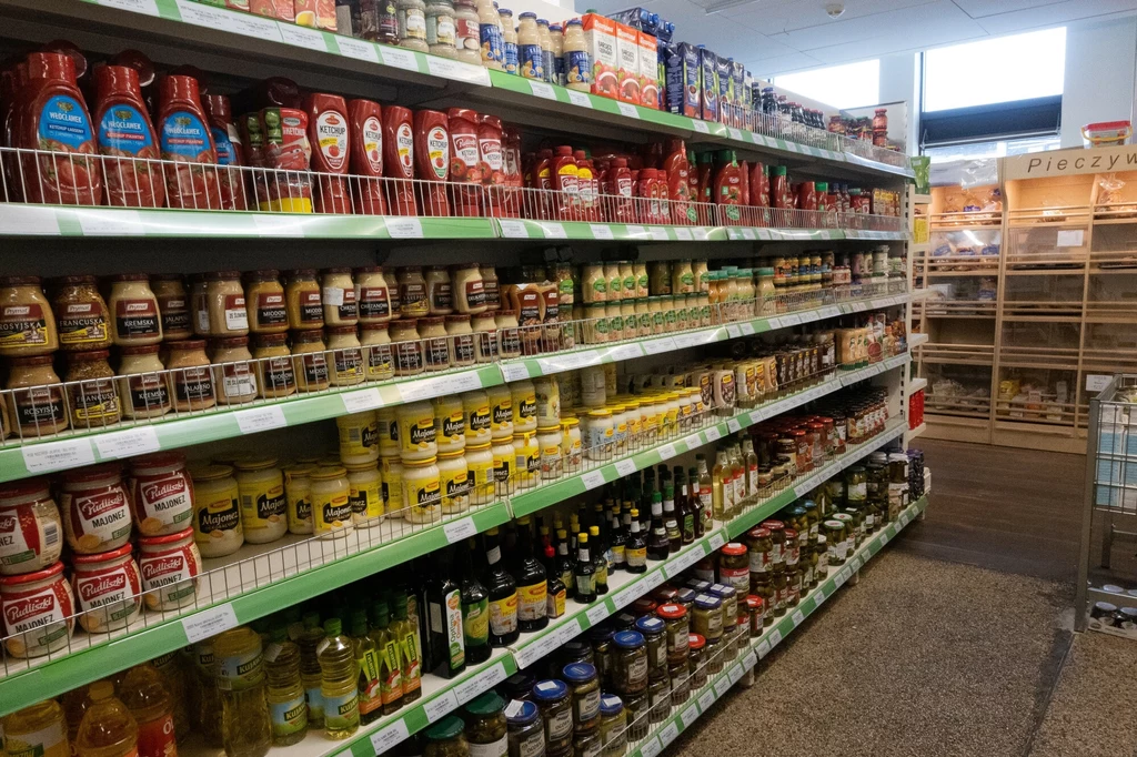 Regały w sklepie spożywczym wypełnione różnymi rodzajami sosów, ketchupów, majonezów, musztard, oliw i innych produktów spożywczych w słoikach i butelkach, z tyłu widoczne stoisko z pieczywem.