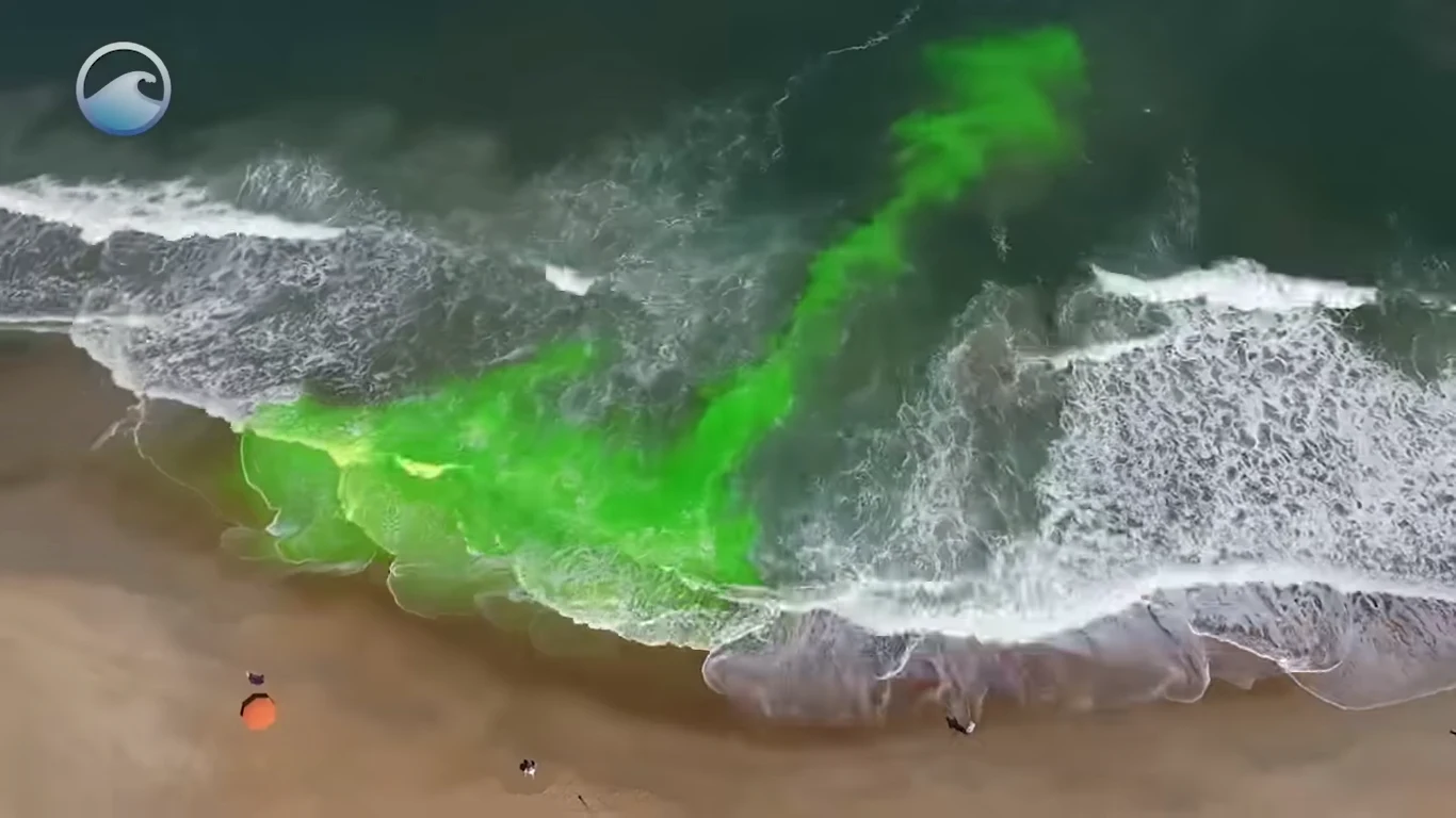 Zielony barwnik rozprzestrzenia się w wodzie przy brzegu oceanu, tworząc wyraźną smugę kontrastującą z naturalnym kolorem fal i piasku na plaży, widoczne są też niewielkie obiekty na plaży, takie jak parasol czy sprzęt plażowy.