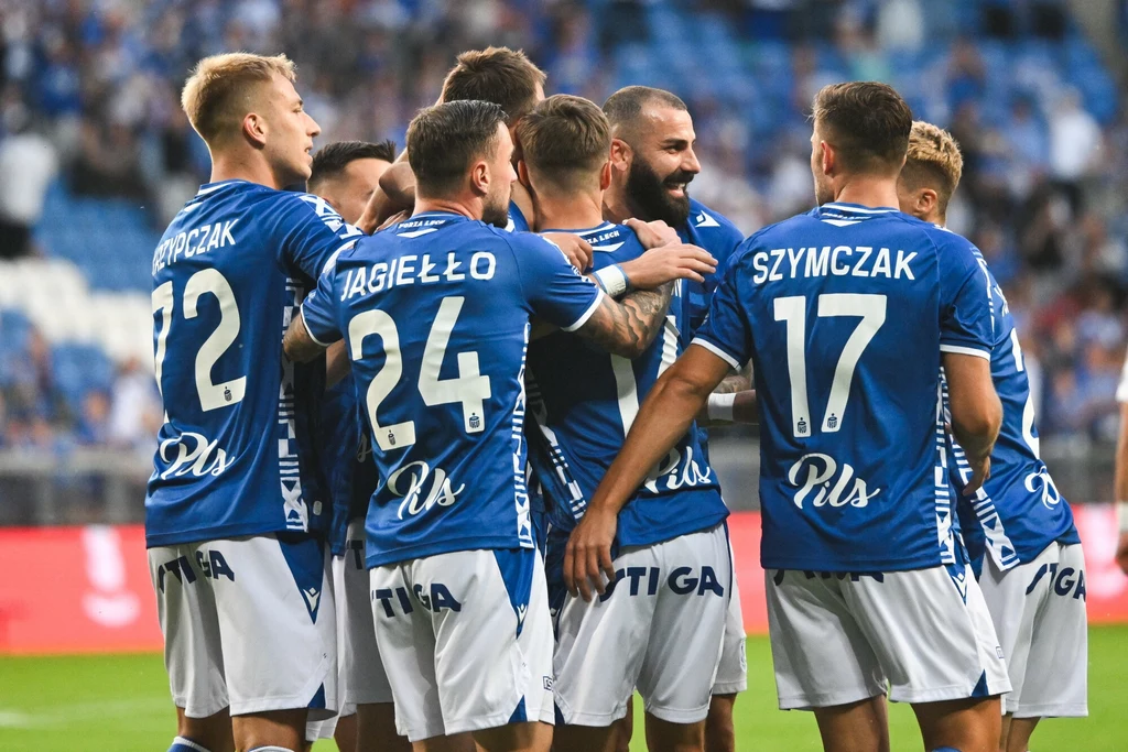 Lech Poznań Grupa piłkarzy w niebieskich koszulkach świętuje wspólnie sukces na boisku, otoczeni są trybunami z kibicami. Na koszulkach widoczne są nazwiska zawodników oraz logotypy sponsorów.