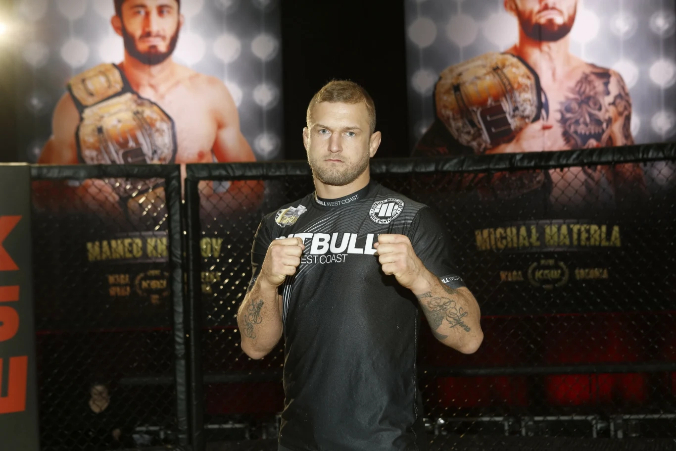 Zawodnik MMA w sportowej odzieży stoi w oktagonie, prezentując gardę gotową do walki. W tle widoczne plakaty innych zawodników z pasami mistrzowskimi.