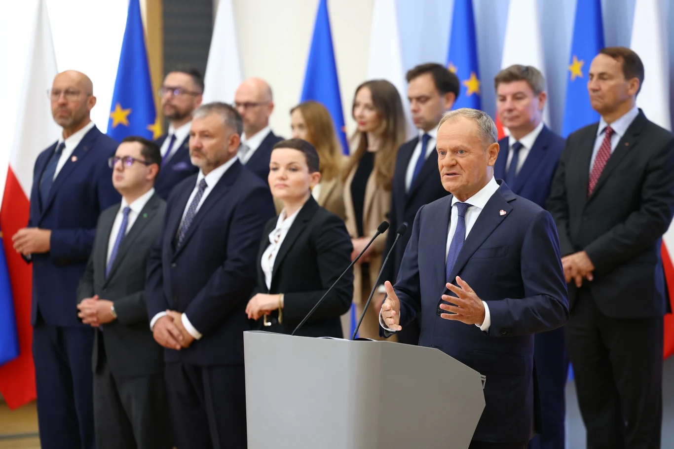 Grupa polityków i urzędników stoi w rzędzie na tle flag Polski i Unii Europejskiej, na pierwszym planie mężczyzna w garniturze przemawia przy mównicy.