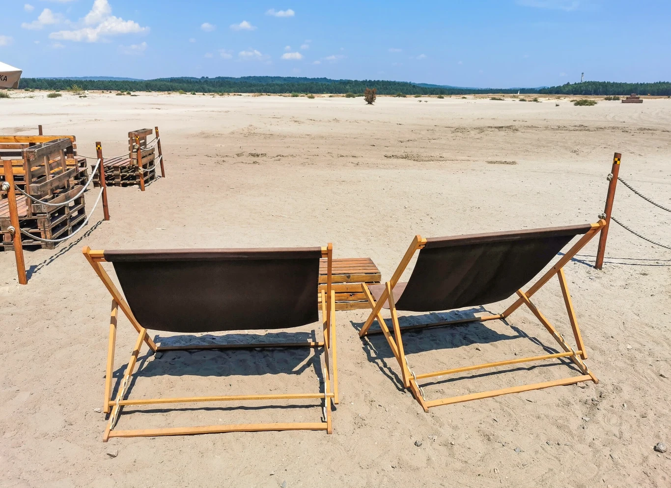 Dwa drewniane leżaki ustawione na piaszczystej plaży z niewielkim drewnianym stolikiem pomiędzy nimi, w tle rozległa przestrzeń piasku oraz niewielka ilość zieleni na horyzoncie.
