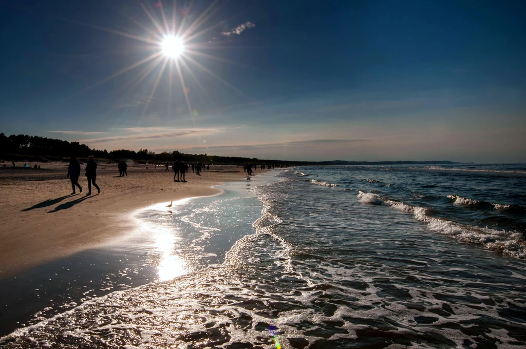 Piaszczysta plaża z falami morskimi, promienie słońca odbijające się na wodzie, sylwetki spacerujących ludzi w oddali, wyraźna linia brzegowa i spokojny, błękitny nieboskłon.