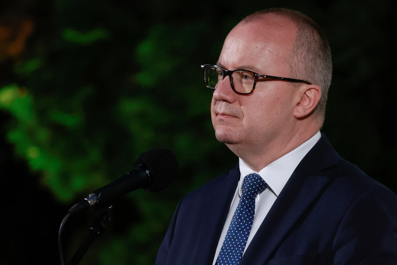 Rekonstrukcja rządu. Adam Bodnar żegna się z Ministerstwem Sprawiedliwości Mężczyzna w okularach i garniturze z krawatem stoi przy mikrofonie na tle rozmytego, zielonego otoczenia.